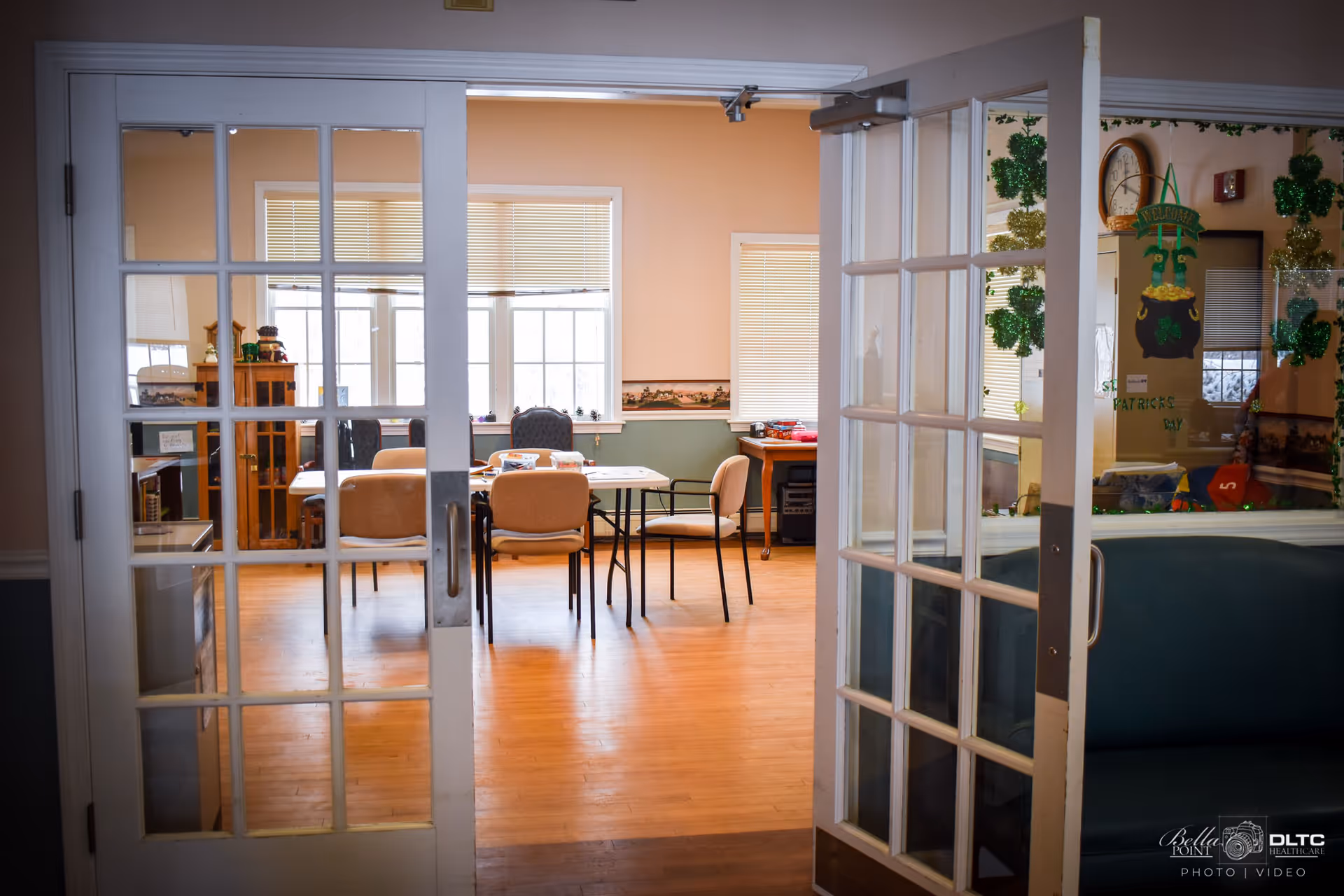 View through open glass-paneled double doors into a room with tables and chairs arranged for activities or meetings, with windows letting in natural light and some decorations on the walls.