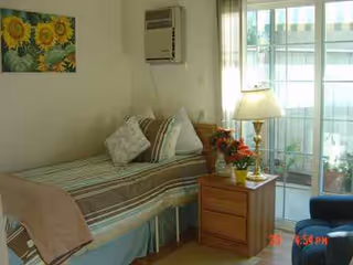 A cozy resident bedroom with bed, nightstand, lamp, flowers, and sliding glass door.