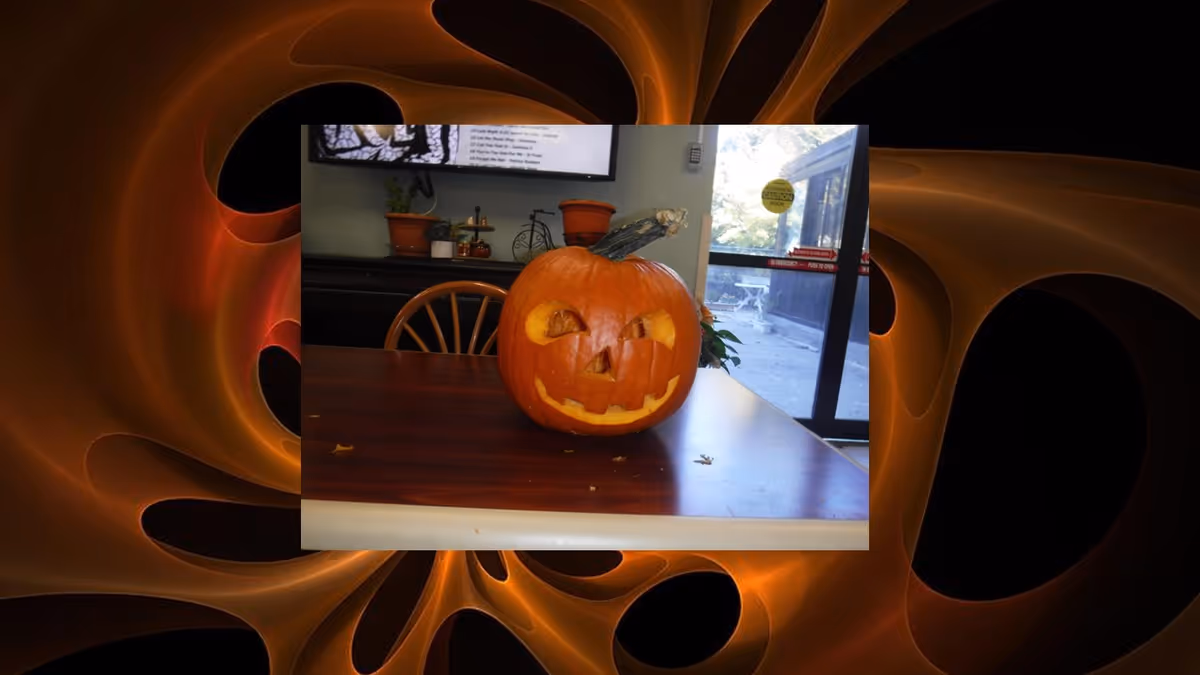 A carved pumpkin with a smiling face placed on a wooden table inside a room. In the background, there is a window showing an outdoor area, a chair, and some potted plants on a shelf.