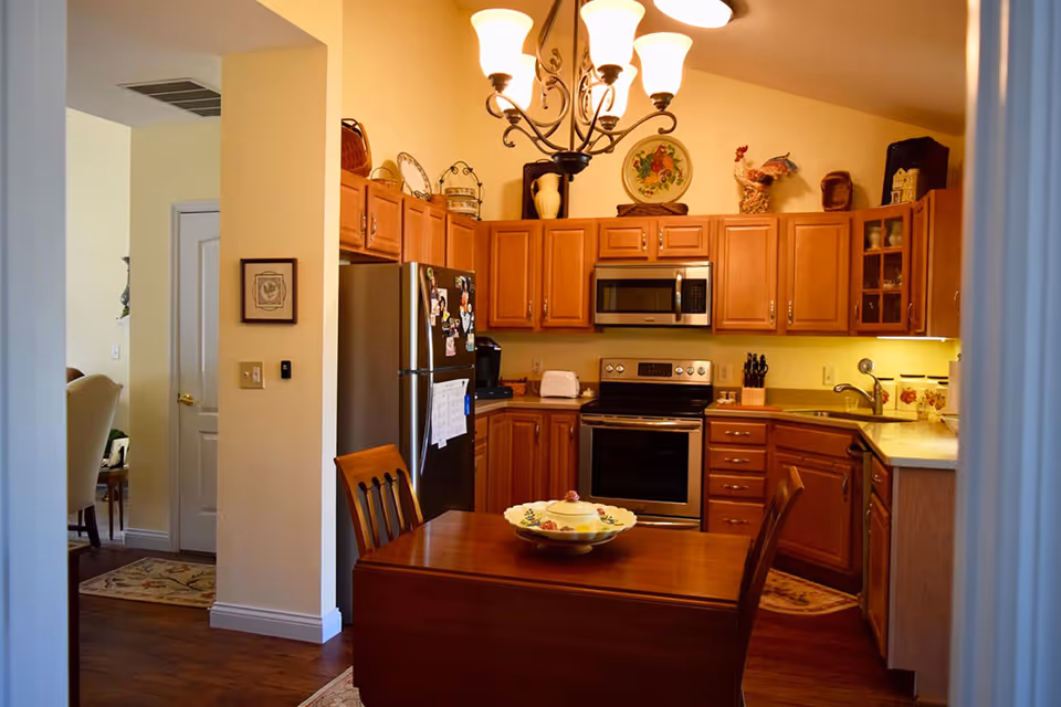 A cozy kitchen with wooden cabinets, stainless steel refrigerator and oven, a microwave above the stove, and a wooden dining table with two chairs. Decorative plates and figurines are displayed on top of the cabinets. Warm lighting from a chandelier illuminates the space.