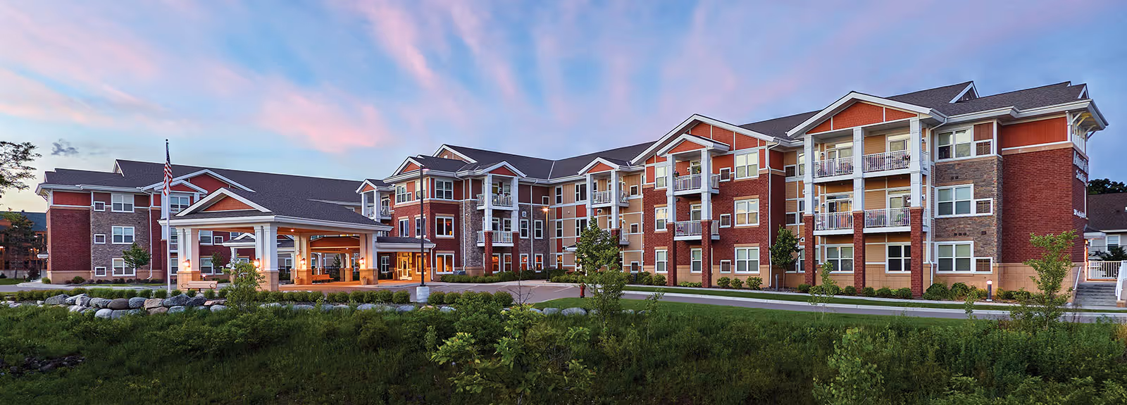 Front exterior of a three-story red-brick senior living building with balconies, a covered entrance, and landscaped lawn under a pastel sky.