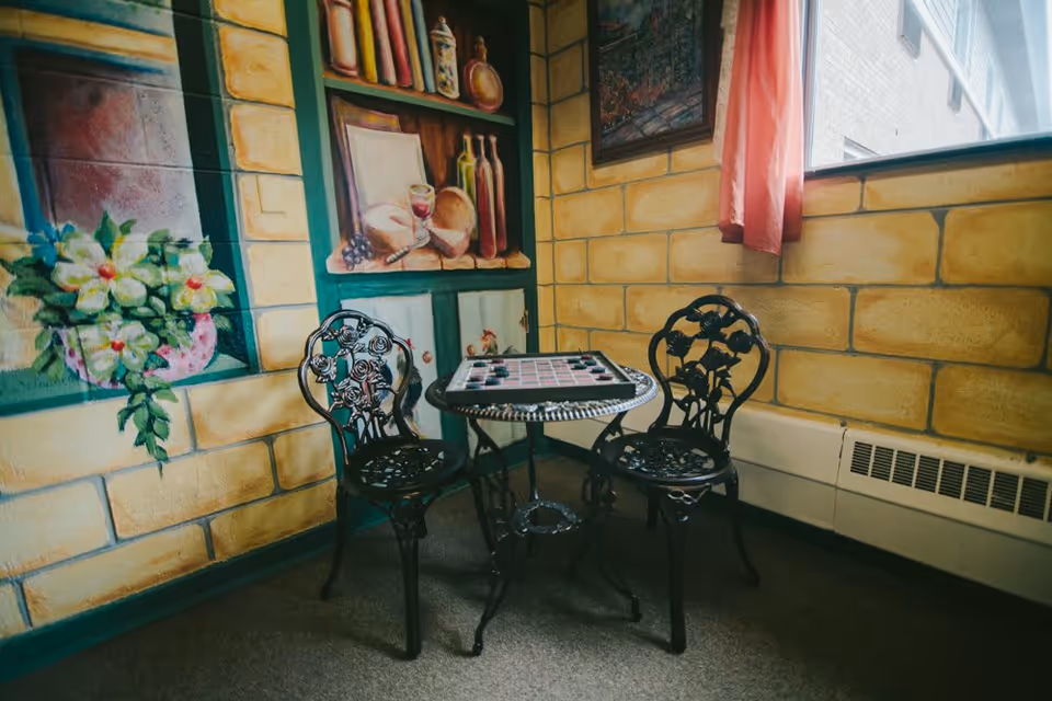 A small corner seating area with two decorative black metal chairs and a round table with a checkerboard on top. The walls are painted with a mural depicting shelves with bottles, bread, and a glass of wine, as well as flowers. A window with a pink curtain lets in natural light.