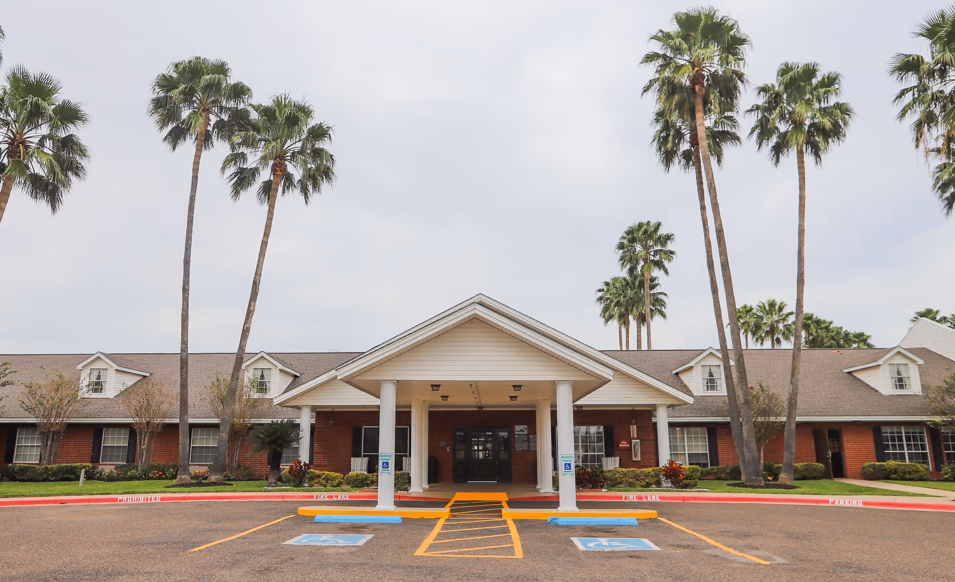 Front entrance of a one-story brick nursing and rehabilitation center with a covered portico, palm trees, and parking spaces.