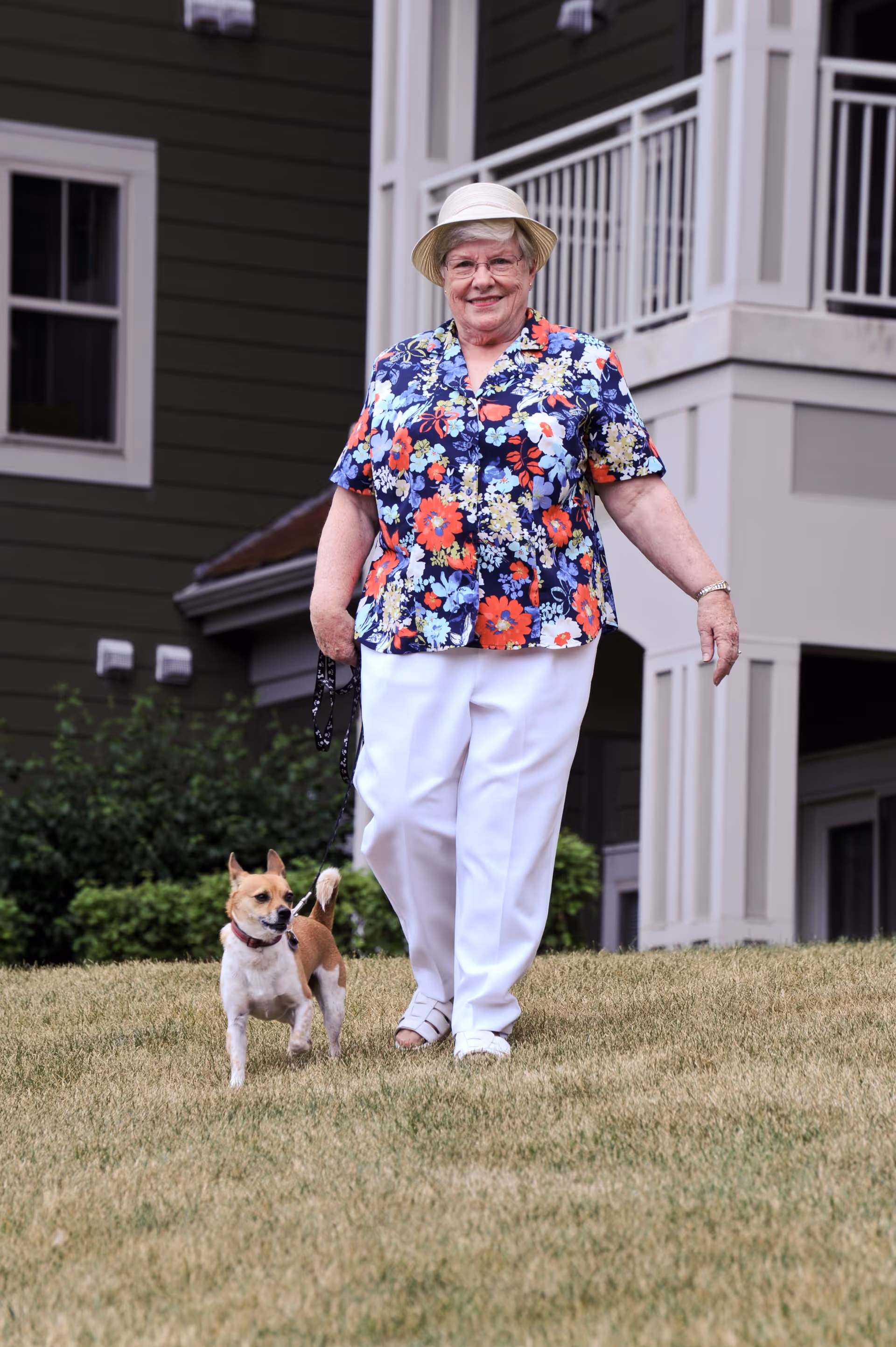 An elderly woman wearing a floral shirt, white pants, and a hat is walking a small dog on a leash outside on a grassy area in front of a residential building.