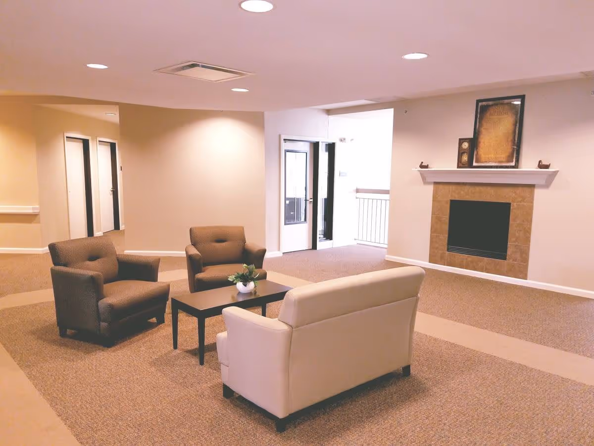 A cozy seating area in a senior living facility with two brown armchairs and a beige loveseat arranged around a small black coffee table with a plant on it. The room has beige walls, carpeted floors, recessed ceiling lights, and a fireplace with decorative items on the mantel. There are doors and a hallway visible in the background.
