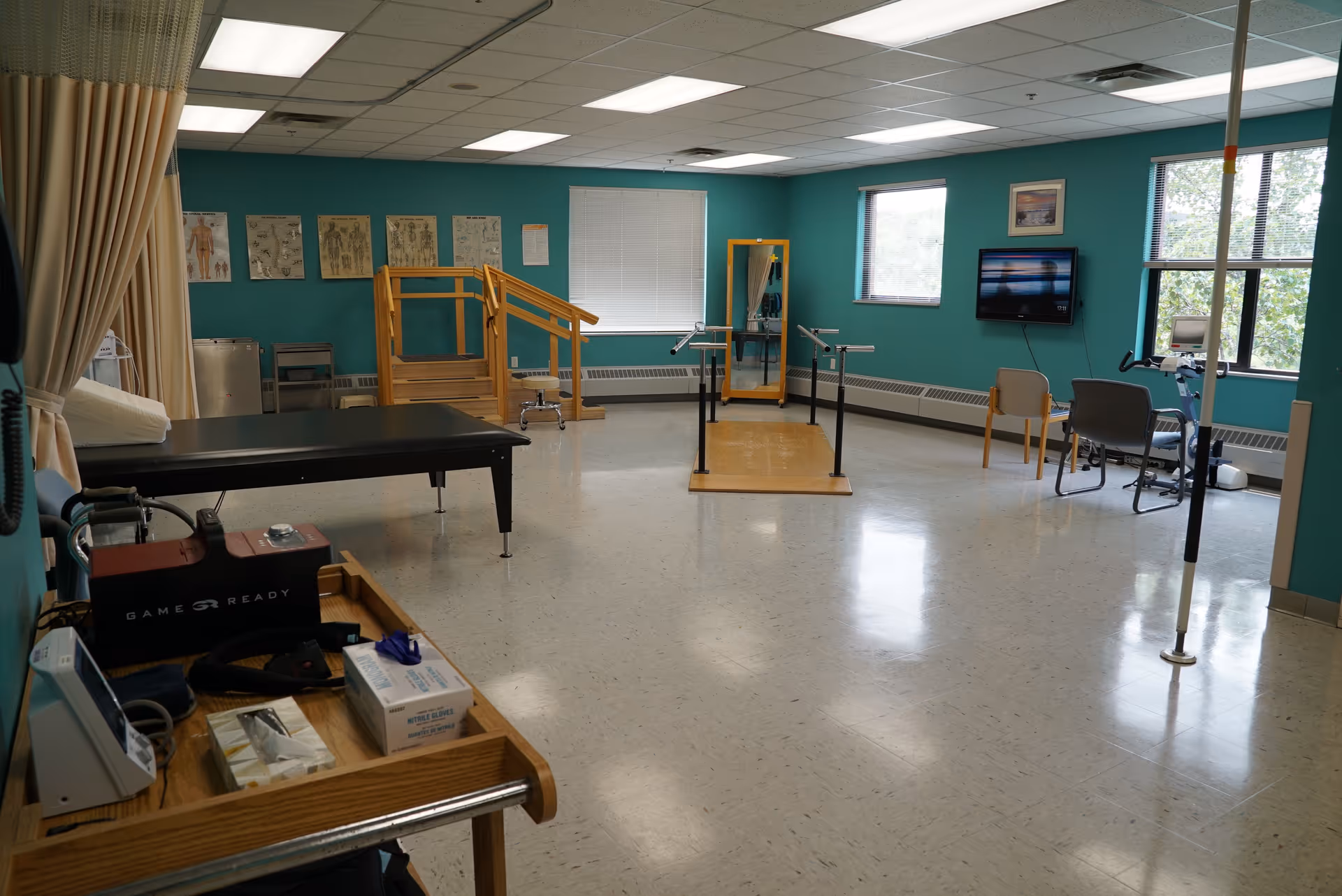 A spacious rehabilitation therapy room with teal walls containing parallel bars, treatment tables, exercise equipment, chairs, and windows.