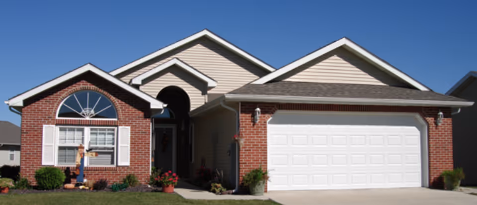 Single-story residential building with a brick and beige siding exterior, a white garage door, and a front entrance with an arched window above it. The house has a well-maintained lawn with some shrubs and potted plants near the entrance.