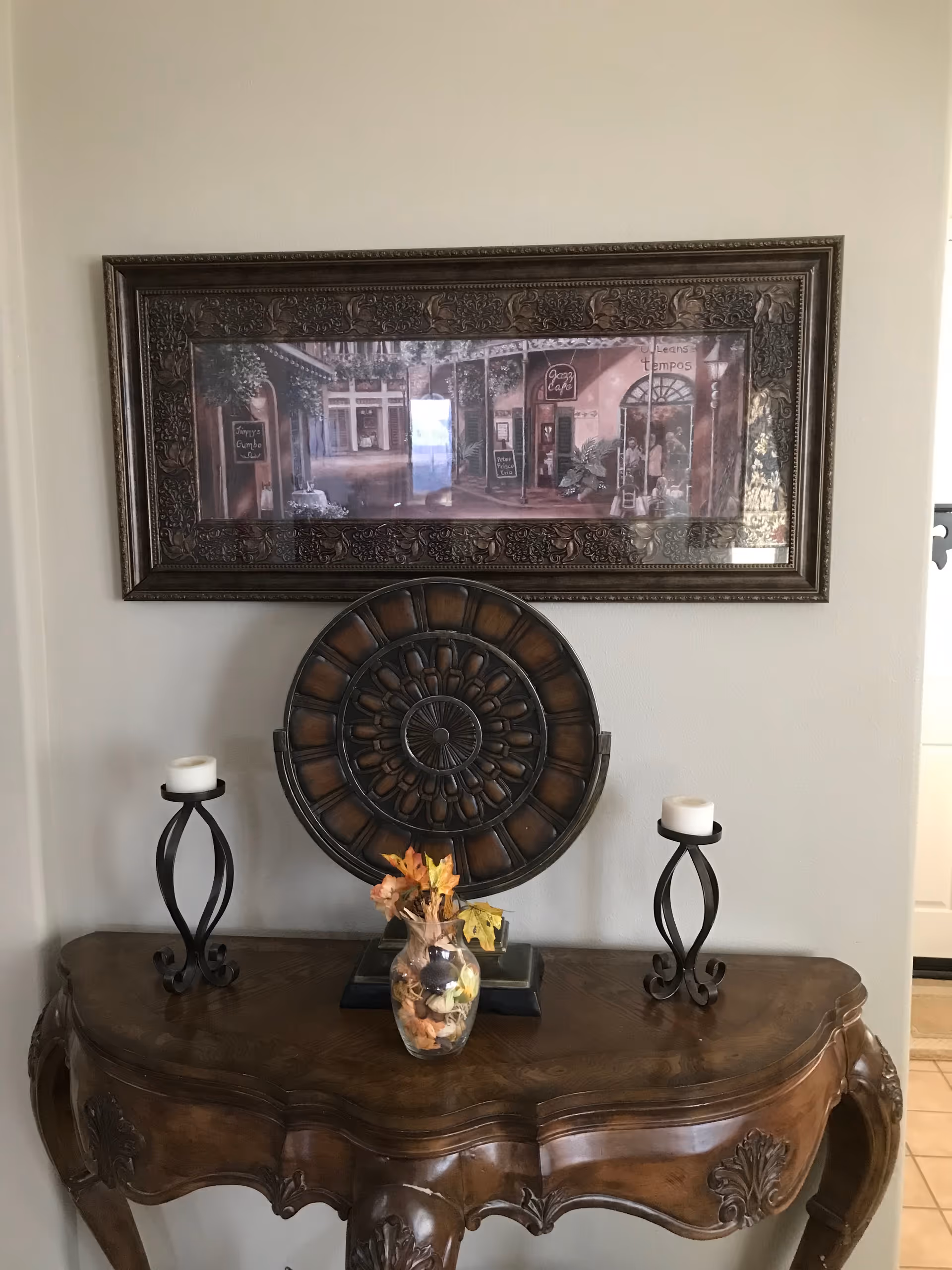A decorative wooden table with intricate carvings holds a glass vase with autumn leaves and two black metal candle holders with white candles. Above the table hangs a large framed painting depicting a street scene with cafes and storefronts.