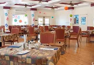 A spacious dining room with multiple tables covered in patterned tablecloths, set with silverware, napkins, and menus. The room has wooden flooring, ceiling fans, and colorful paper decorations hanging from the ceiling and walls. Large windows and double doors provide natural light.
