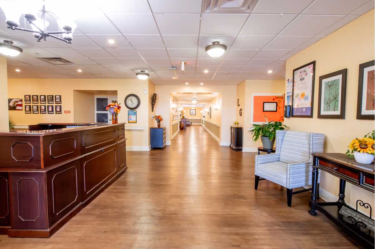 Bright senior living facility reception area with a wooden front desk, seating, framed art, and a long central hallway.