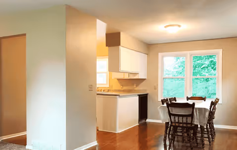 Open-plan dining area with a wooden table and chairs by a window adjacent to a small kitchen.