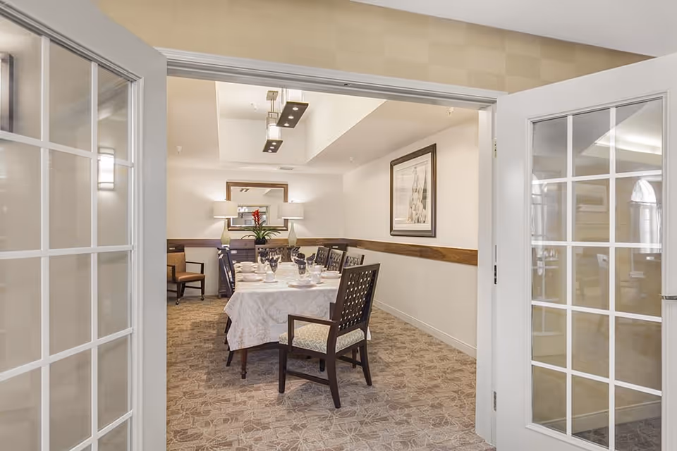 View through open double glass doors into a dining room with a long table covered with a white tablecloth, set with plates, glasses, and napkins. The room has carpeted floors, a framed artwork on the wall, two table lamps on a sideboard, and a ceiling light fixture.
