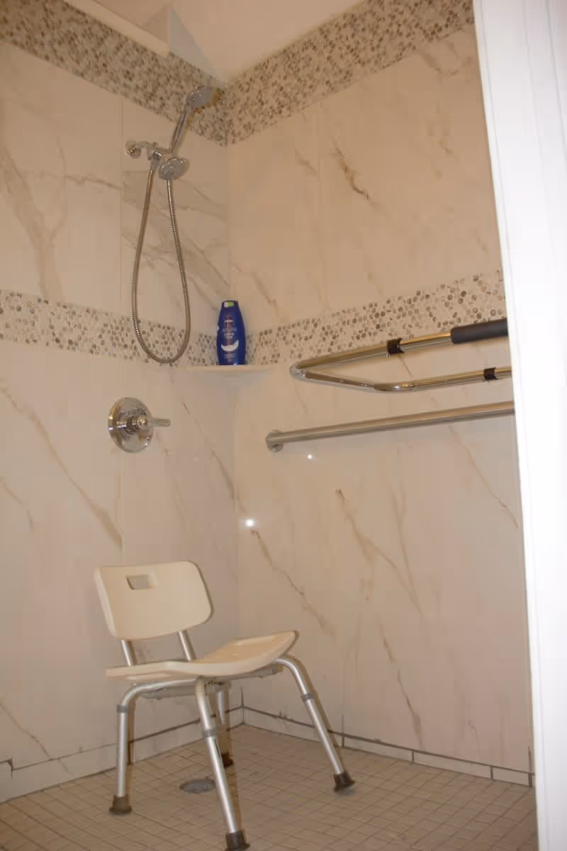 Shower area with beige marble-patterned tiles and a decorative mosaic tile border. There is a handheld showerhead attached to the wall, a white shower chair with metal legs, a grab bar, and a bottle of body wash on a corner shelf.