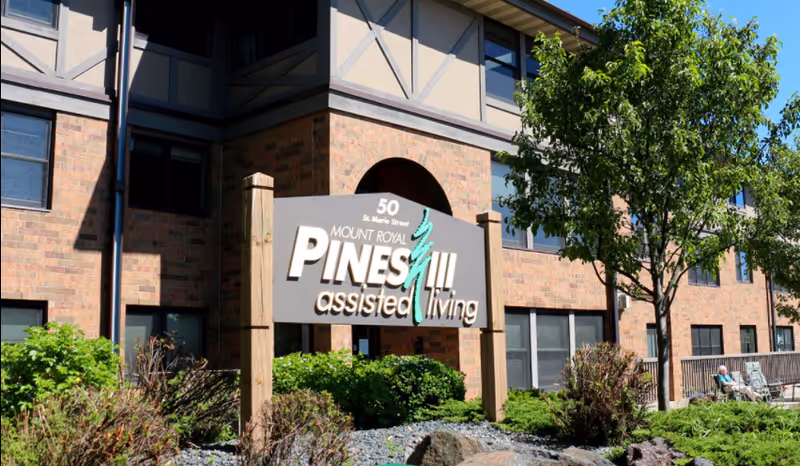 Sign reading 'PINES assisted living' in front of a brick multi-story senior living building with landscaping and a tree.