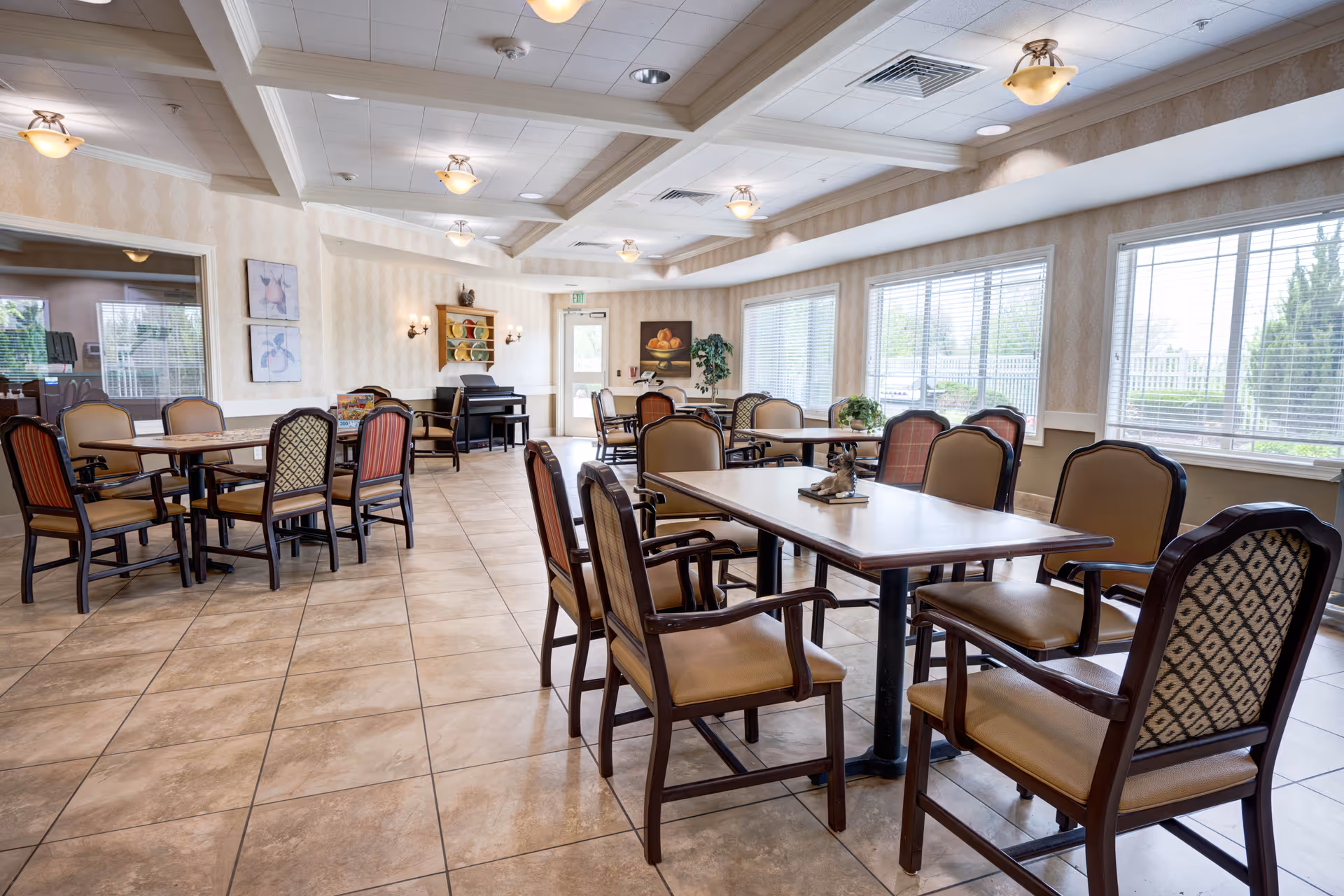 A bright and spacious dining room with multiple tables and cushioned chairs arranged neatly. Large windows on the right side allow natural light to fill the room. The ceiling has recessed lighting and decorative fixtures. The walls are adorned with framed artwork and a small shelf with decorative plates. The floor is tiled with a light brown pattern.