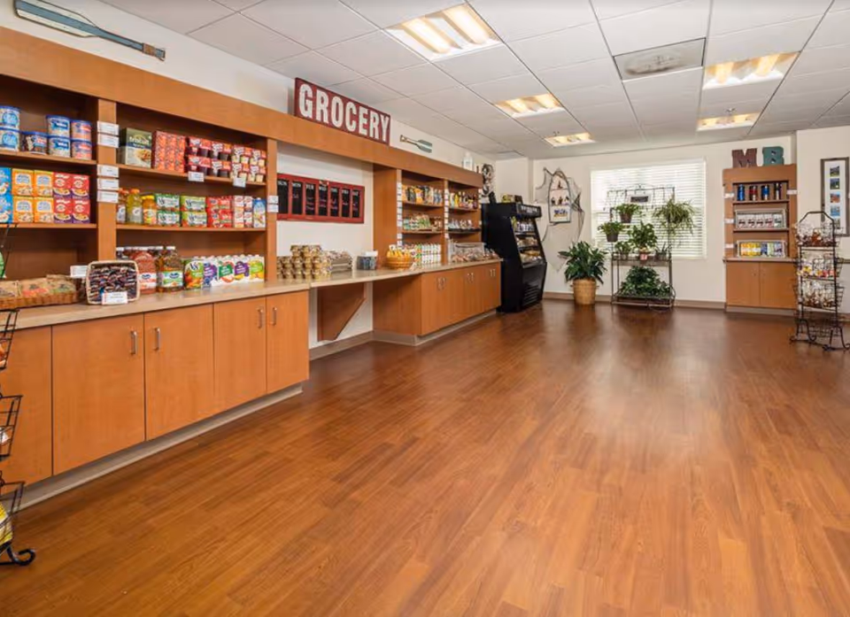 Interior view of a small grocery area in a senior living facility with wooden shelves stocked with various food items and snacks. The room has wood flooring, a large window with plants on a metal rack, and bright ceiling lights.
