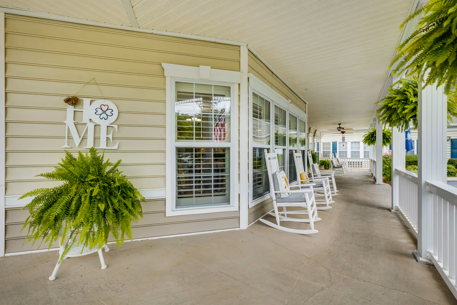Covered front porch of a senior living building with white rocking chairs, hanging ferns, and a decorative 'HOME' sign on beige siding.