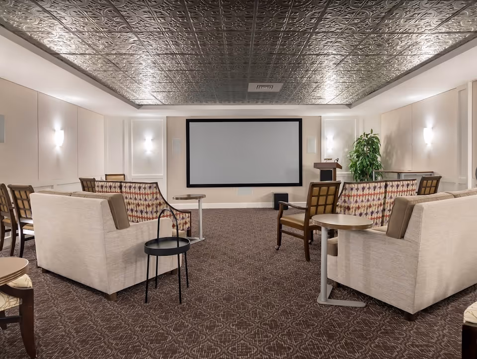 A senior living facility media room with a large blank projector screen on the front wall. The room has patterned carpet, beige walls with wall sconces, and a decorative metallic ceiling. There are several cushioned chairs and sofas arranged facing the screen, along with small side tables and a podium with a microphone to the right of the screen. A potted plant is placed in the corner near the podium.