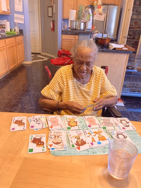 An elderly woman wearing a yellow and white striped shirt is sitting at a wooden table in a kitchen or dining area. She is playing a card game with colorful animal-themed cards spread out in front of her. The background shows kitchen cabinets, a refrigerator, and various kitchen items.