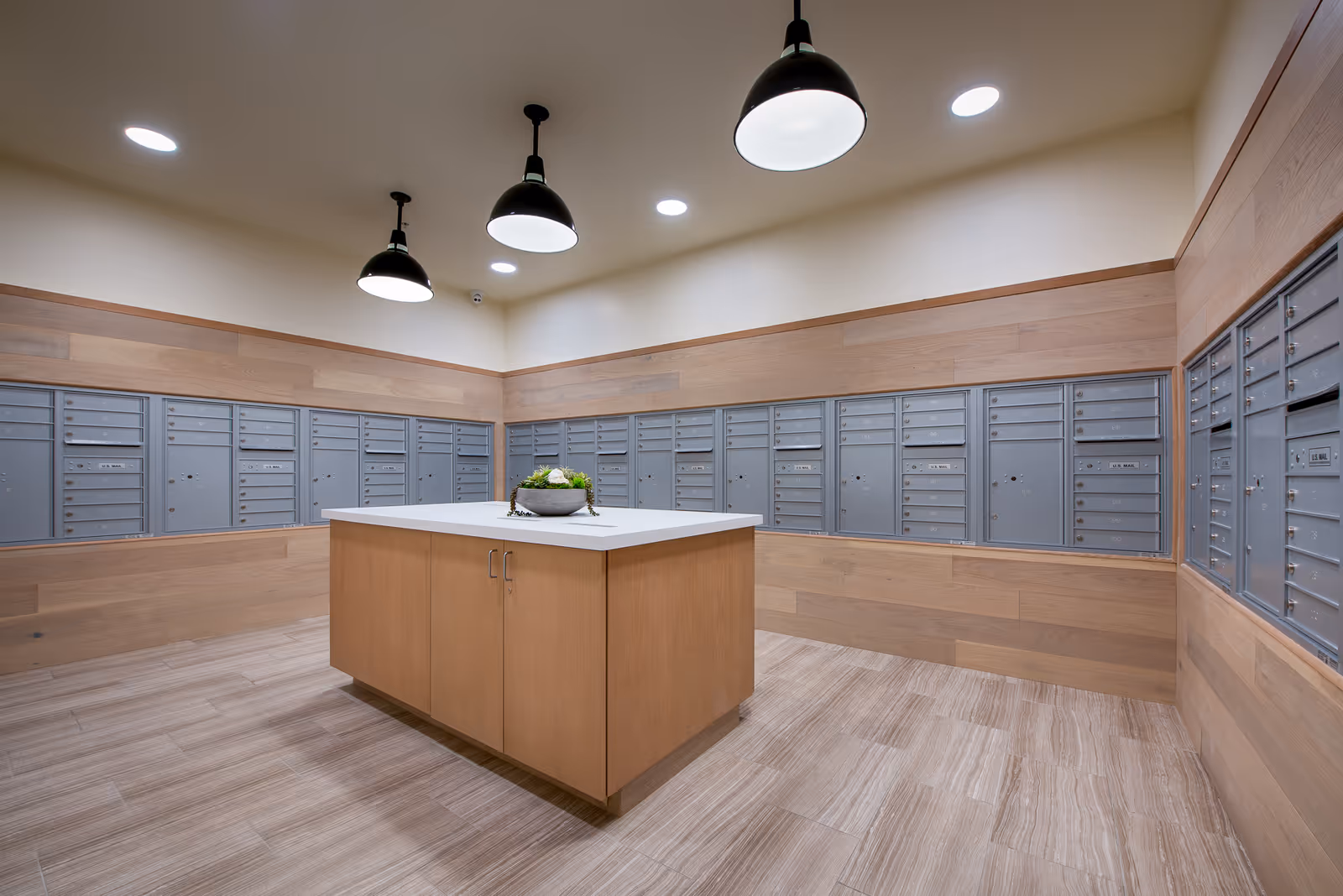 Interior room with multiple gray mailboxes mounted on light wood-paneled walls, a central wooden island with a white countertop and a small decorative plant, and three black pendant lights hanging from the ceiling.