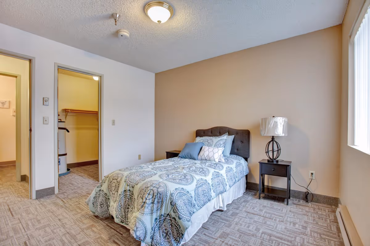 A small bedroom with a single bed covered in a blue and white patterned comforter and multiple pillows. There is a dark wooden nightstand with a lamp on the right side of the bed. The room has beige walls and carpeted floor. Two open doorways are visible on the left side, one leading to a closet with a water heater and the other to another room.