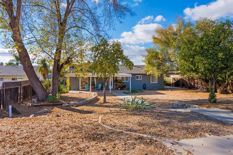 Single-story gray house with a covered patio and chairs, a dry landscaped backyard with trees and a curving concrete path under a blue sky.