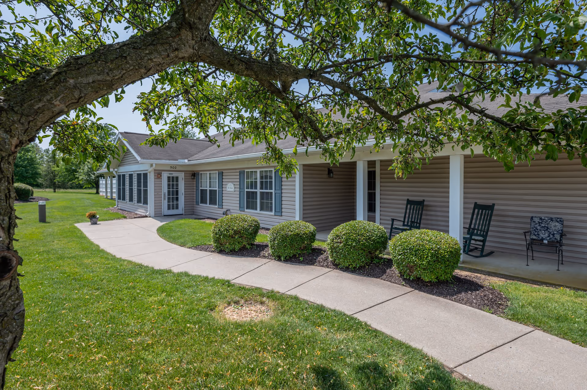 Exterior view of a single-story senior living community building with beige siding, white trim, and a covered porch area with rocking chairs. A concrete walkway curves through a well-maintained lawn with bushes and a tree with green leaves in the foreground.