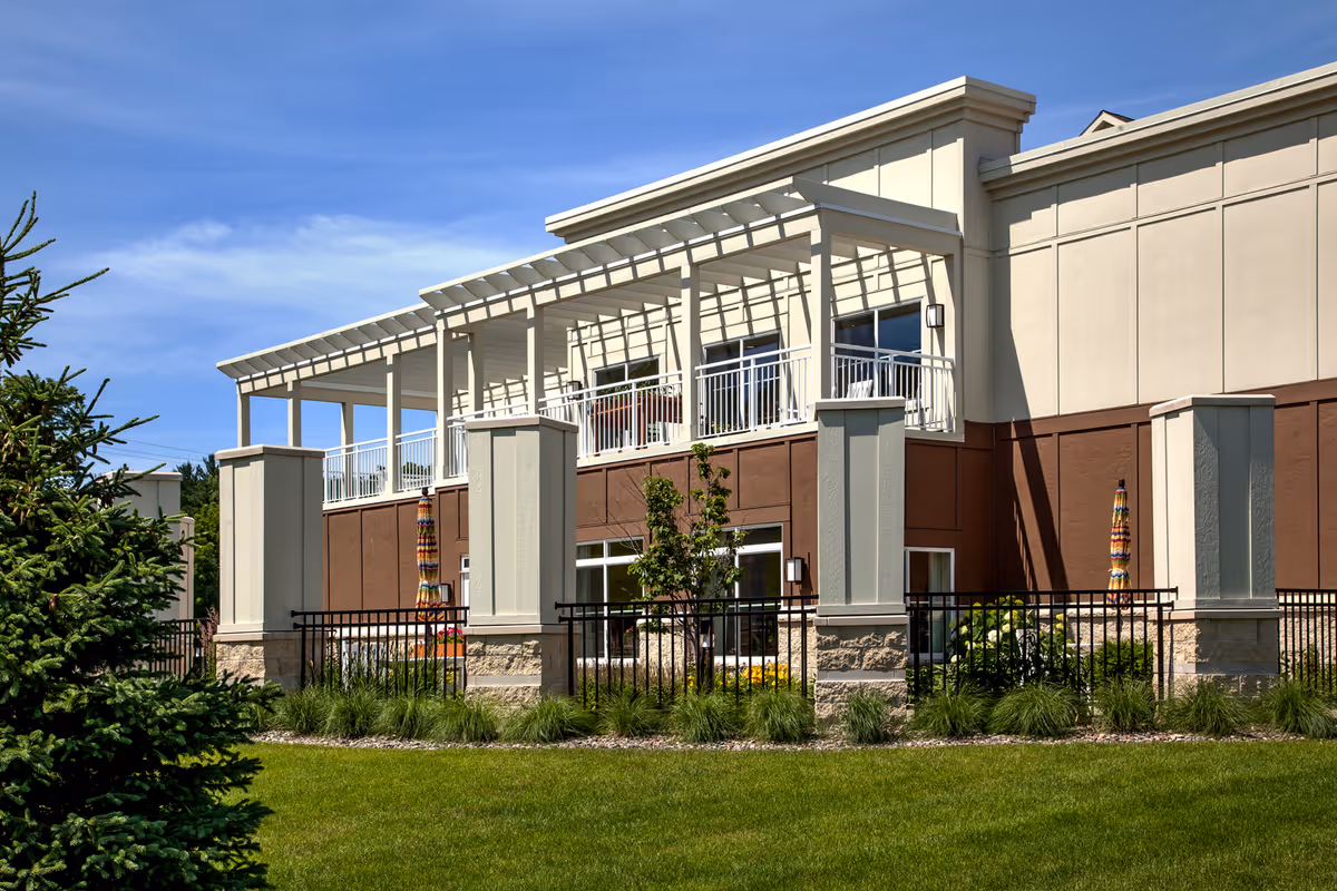 Exterior view of a senior living facility building with a beige and brown facade, featuring a balcony with white railings and a pergola. The building is surrounded by a black metal fence, green grass, shrubs, and a tree, under a clear blue sky.