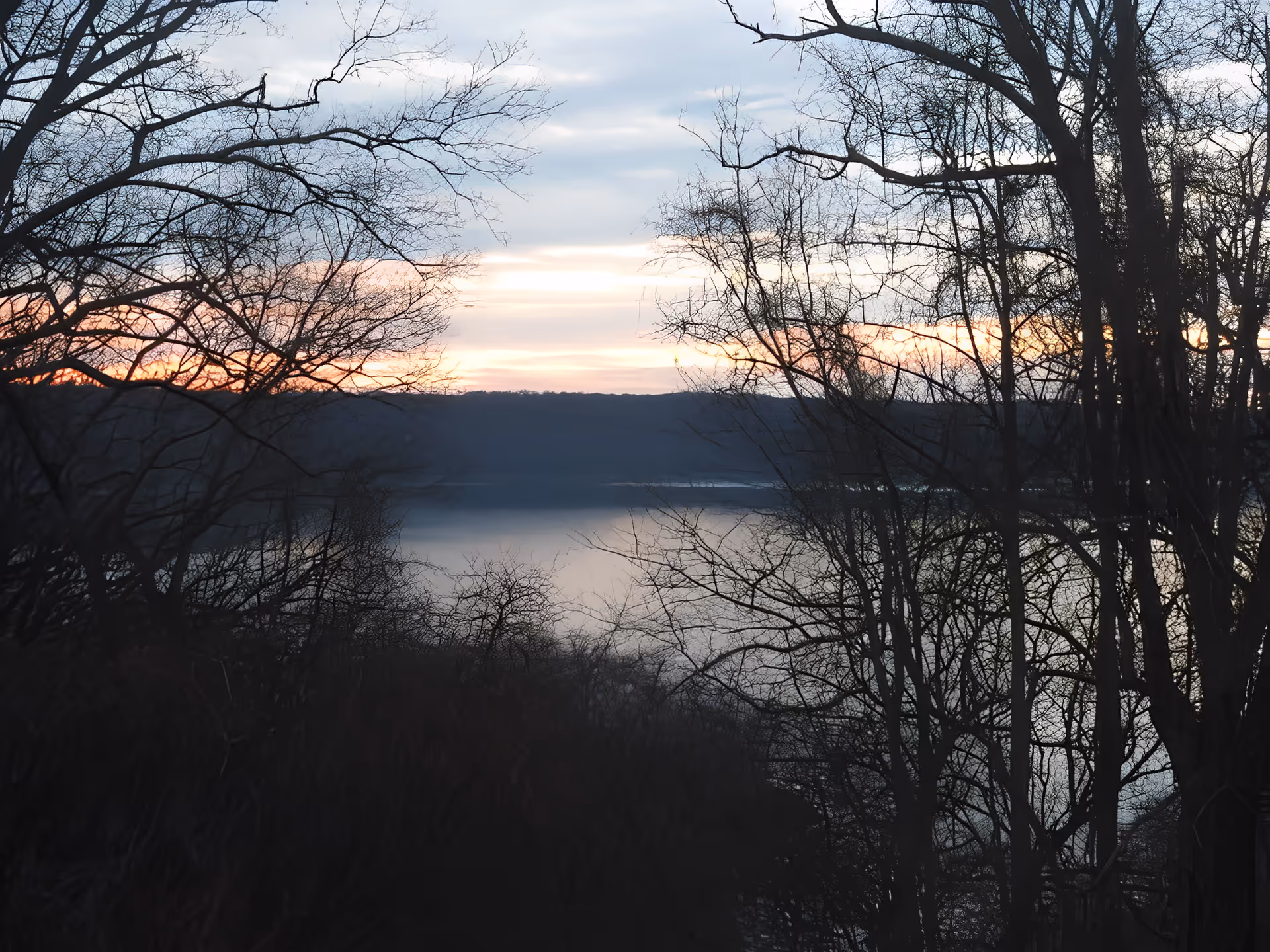 View of a calm lake at sunset with bare trees in the foreground and hills in the background under a partly cloudy sky.