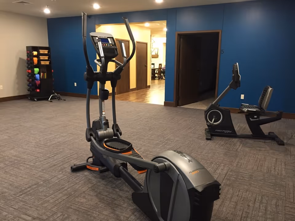 Interior view of a fitness room with exercise equipment including an elliptical machine and a recumbent bike. There is a rack with colorful dumbbells and exercise mats against a blue wall. The room has carpeted flooring and an open doorway leading to a hallway with wooden floors and additional rooms.