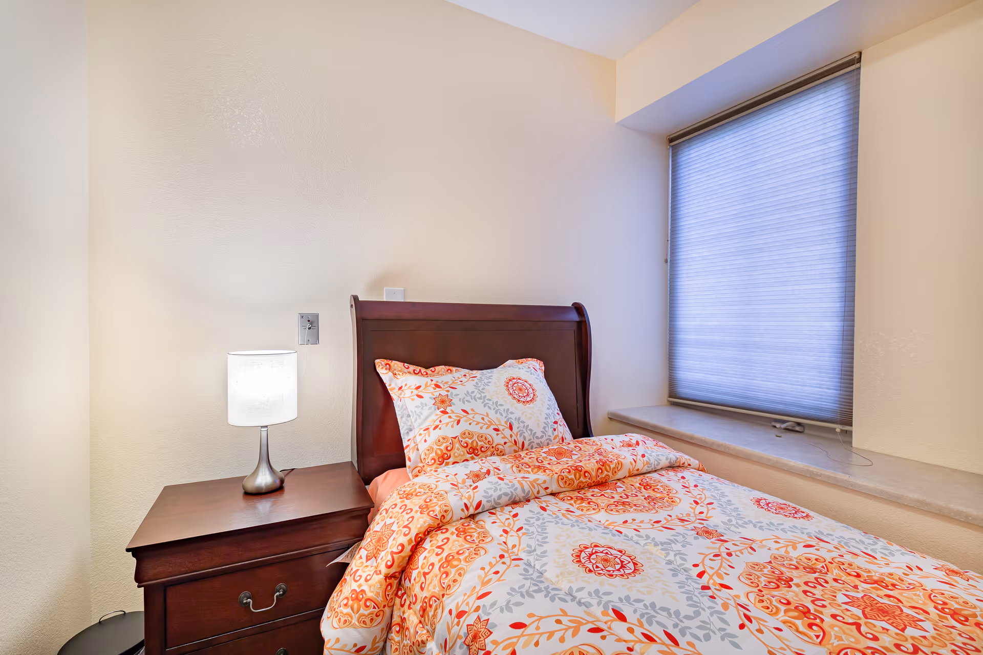 A cozy bedroom featuring a single wooden bed with a colorful floral-patterned comforter and pillow in shades of orange, red, and gray. Next to the bed is a wooden nightstand with a modern silver lamp emitting soft light. A window with a closed blue blind is situated above a wide windowsill on the right side of the bed. The walls are painted a light cream color.
