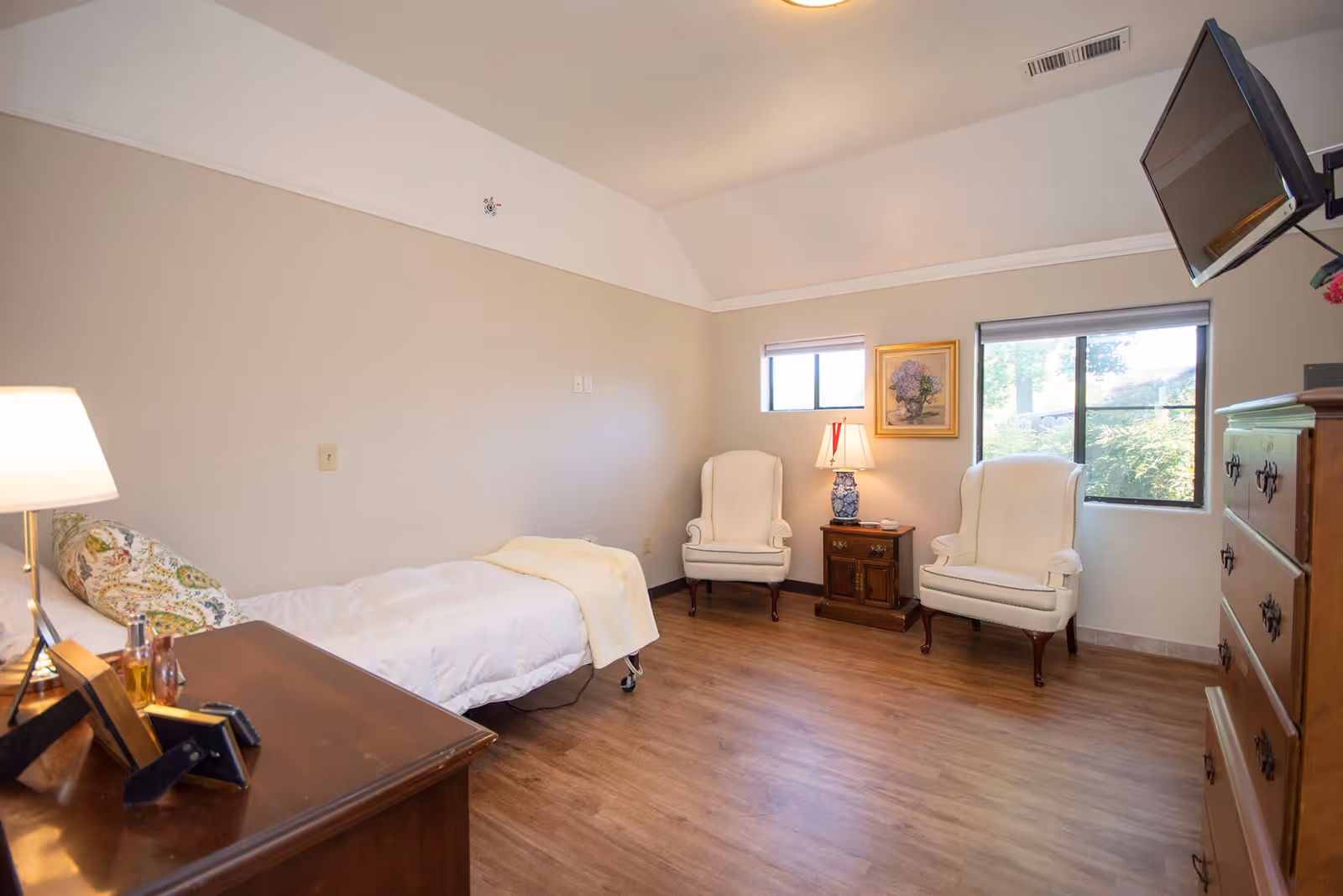 A bright and cozy bedroom with a single bed covered in white bedding and a patterned pillow. There is a wooden dresser with a lamp and picture frames on it. Two white upholstered armchairs are placed near two windows, one small and one larger, letting in natural light. A wooden nightstand with a blue and white lamp and a framed floral painting hangs on the wall. A flat-screen TV is mounted on the wall above a wooden chest of drawers.