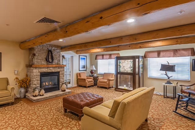 Cozy living room area with a stone fireplace, wooden ceiling beams, patterned carpet, beige sofas, two armchairs, a brown tufted ottoman, a rocking chair, and large windows with blinds and valances.