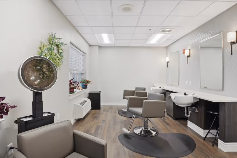 Interior view of a salon area in a senior living facility featuring two salon chairs with footrests, two sinks with mirrors above them, a hair dryer hood, potted plants on the windowsill, and wood flooring.