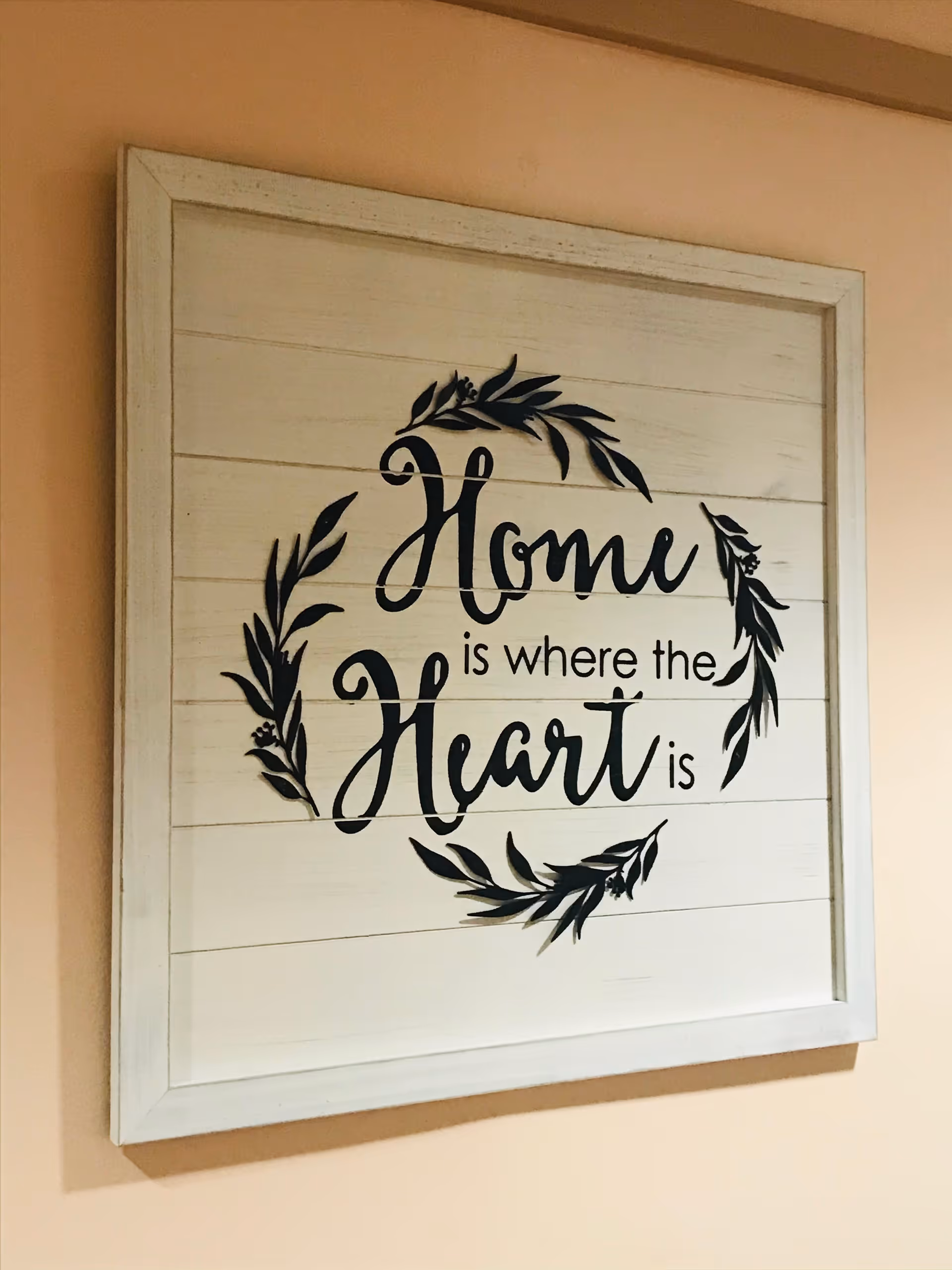 A framed wall decoration with the phrase 'Home is where the Heart is' written in black cursive and regular fonts, surrounded by a circular leafy design, hanging on a peach-colored wall.