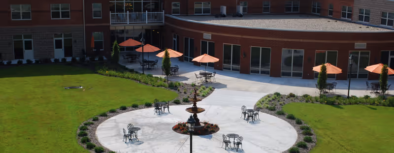 Outdoor courtyard area of Paramount Senior Living Bethel Park featuring a circular concrete patio with a central fountain, surrounded by metal tables and chairs. Several orange umbrellas provide shade over some seating areas. The courtyard is bordered by well-maintained grass and landscaping, with the senior living facility building visible in the background.
