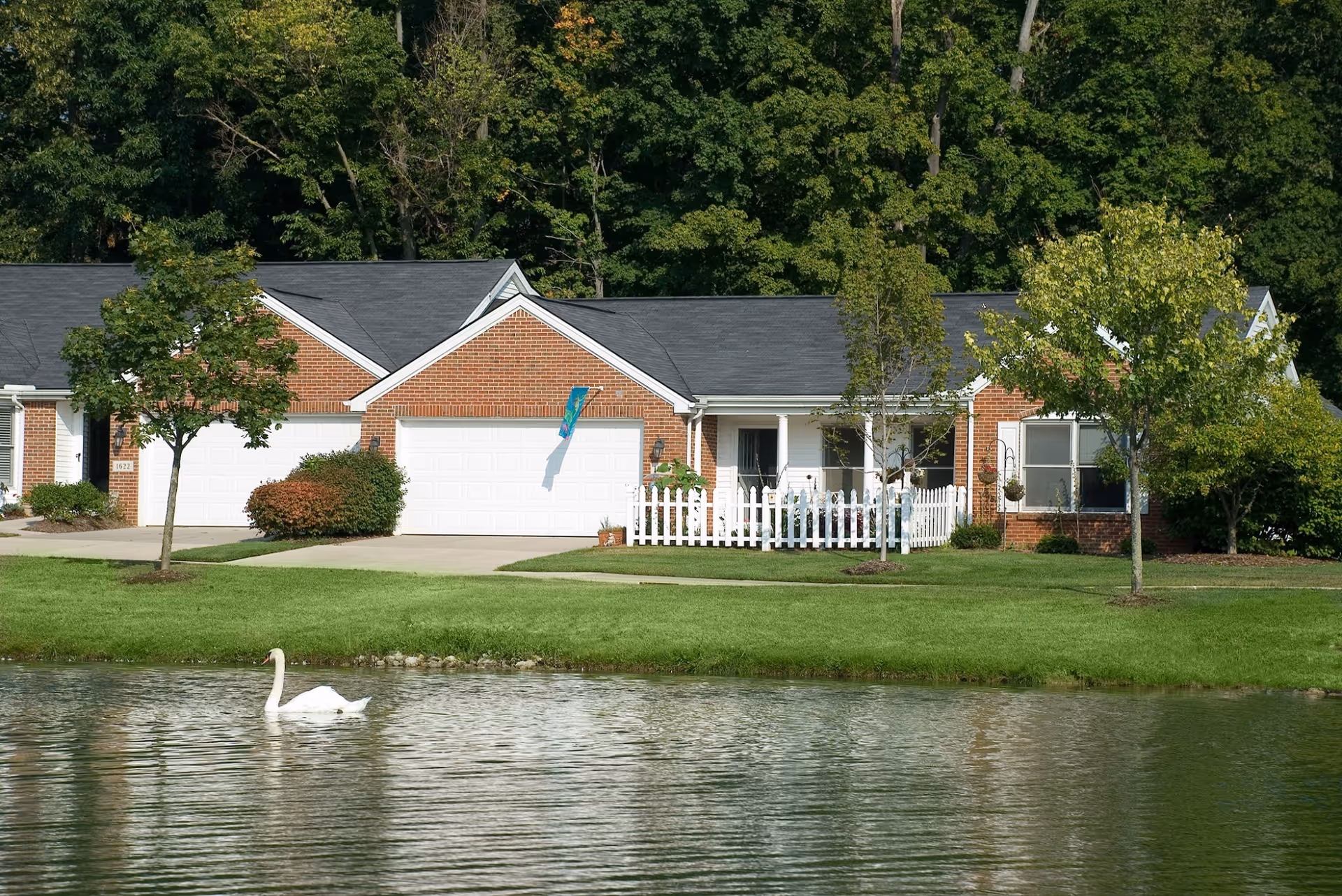 Brick ranch-style residential units with garages and a white picket fence overlooking a pond where a swan is swimming.