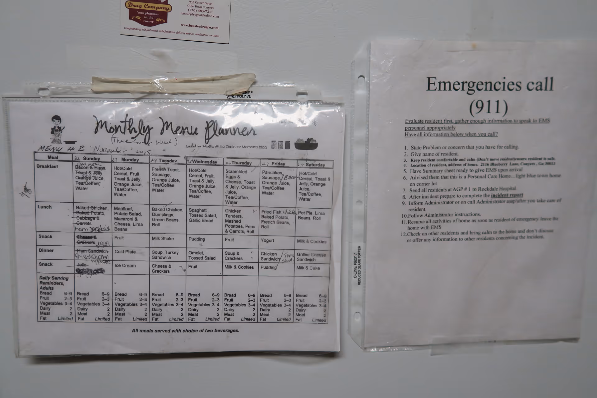 Two laminated sheets attached to a white refrigerator. The left sheet is a monthly menu planner for November 2015 with meals listed for each day of the week, including breakfast, lunch, snack, and dinner options. The right sheet provides emergency instructions for calling 911, detailing steps to evaluate the resident, gather information, and follow up after the call.