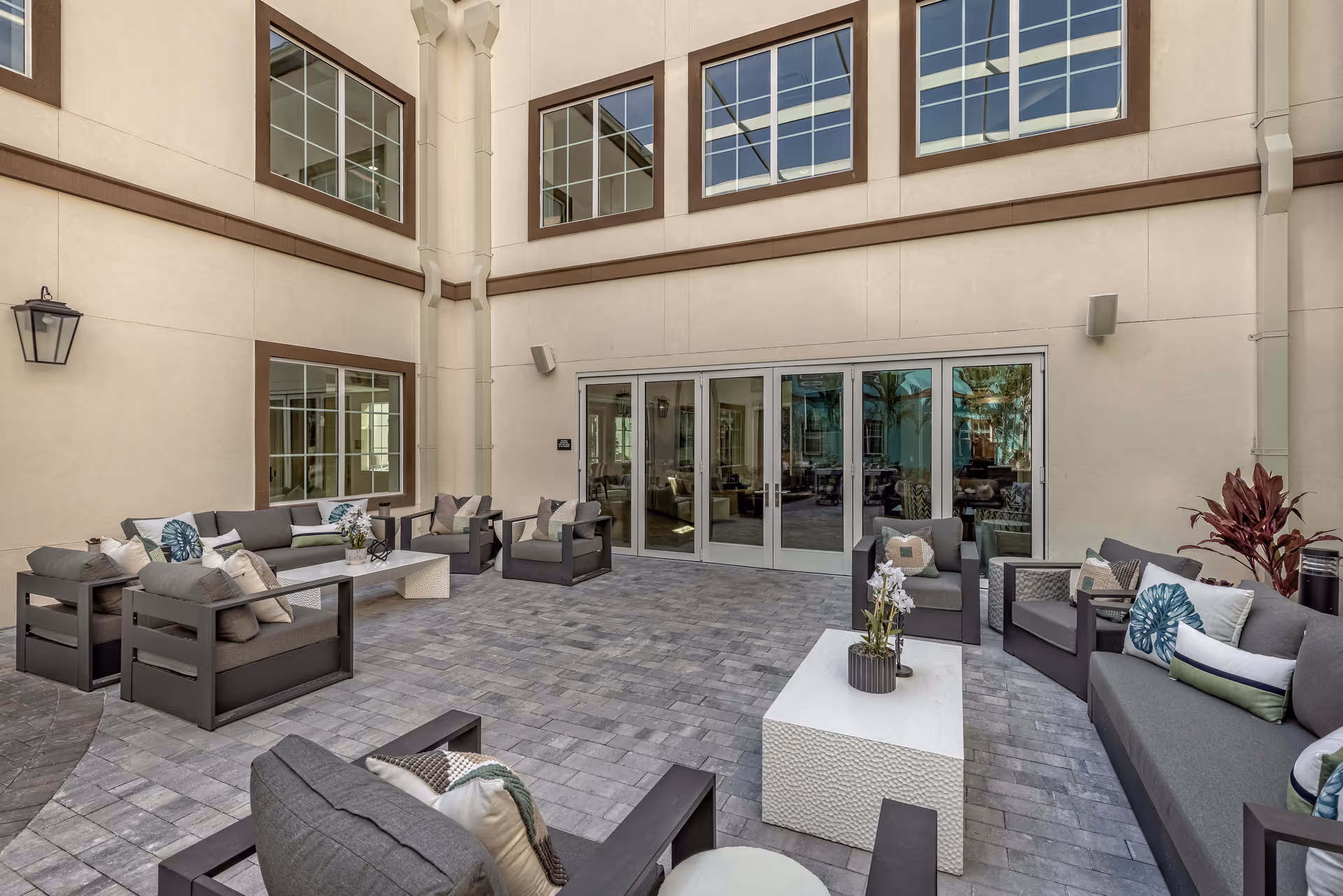 Outdoor courtyard area with modern patio furniture including sofas and armchairs arranged around white coffee tables. The space is enclosed by beige walls with large windows and glass doors leading inside. The floor is paved with gray stone tiles, and there are decorative plants on the tables and in the corner.