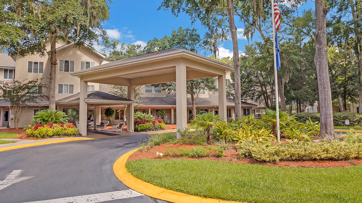 Covered porte-cochere at the entrance of a multi-story senior living building with landscaped grounds and flagpoles.