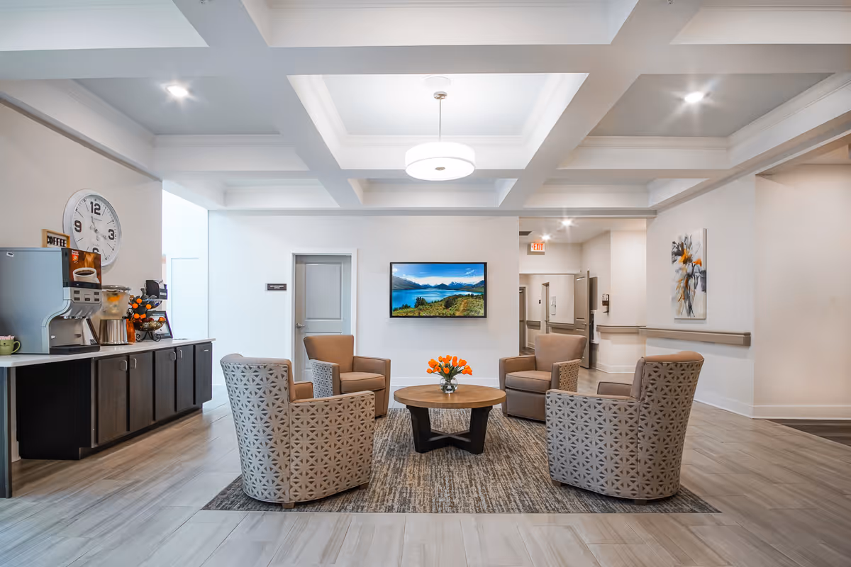 A bright and modern senior living facility common area with four cushioned armchairs arranged around a round wooden coffee table with a vase of orange flowers. A flat-screen TV is mounted on the wall displaying a scenic landscape. To the left, there is a coffee station with a coffee machine, cups, and a large wall clock. The room has a coffered ceiling with a central light fixture and light-colored flooring.