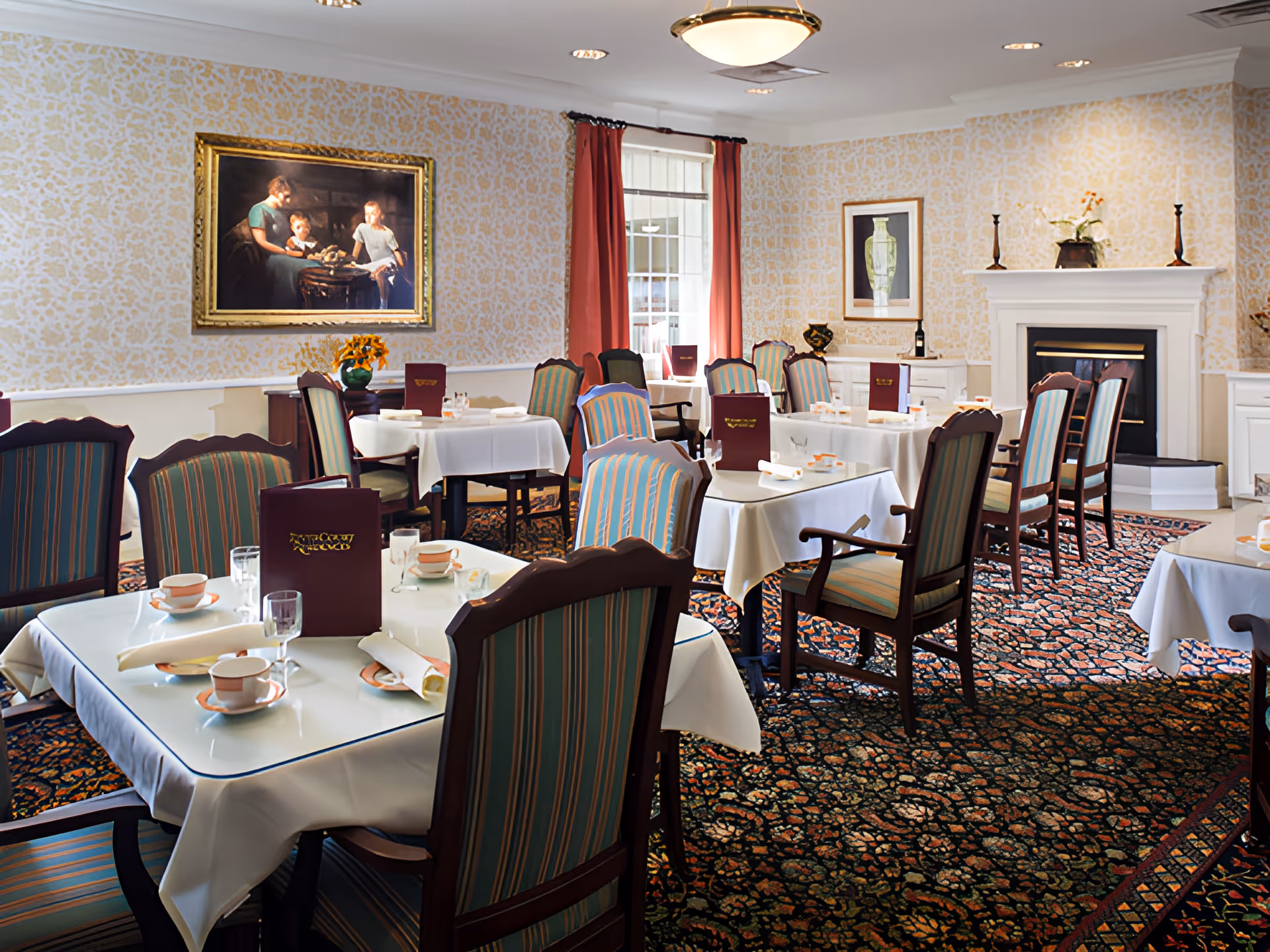 A dining room with multiple tables covered in white tablecloths, each set with cups, saucers, glasses, and rolled napkins. The chairs have striped upholstery. The room features patterned wallpaper, a carpet with a floral design, a fireplace, framed artwork on the walls, and red curtains on the window.