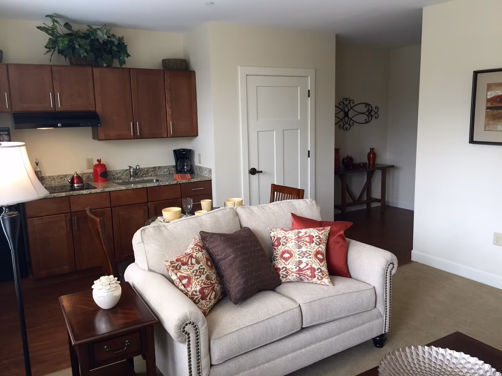 A cozy living area with a beige loveseat adorned with patterned and solid pillows. Behind the loveseat is a small kitchen area with wooden cabinets, a granite countertop, a coffee maker, and a red kettle. A wooden side table with a decorative white flower vase and a floor lamp are also visible. The walls are light-colored, and there is a door and a small table with decorative items in the background.