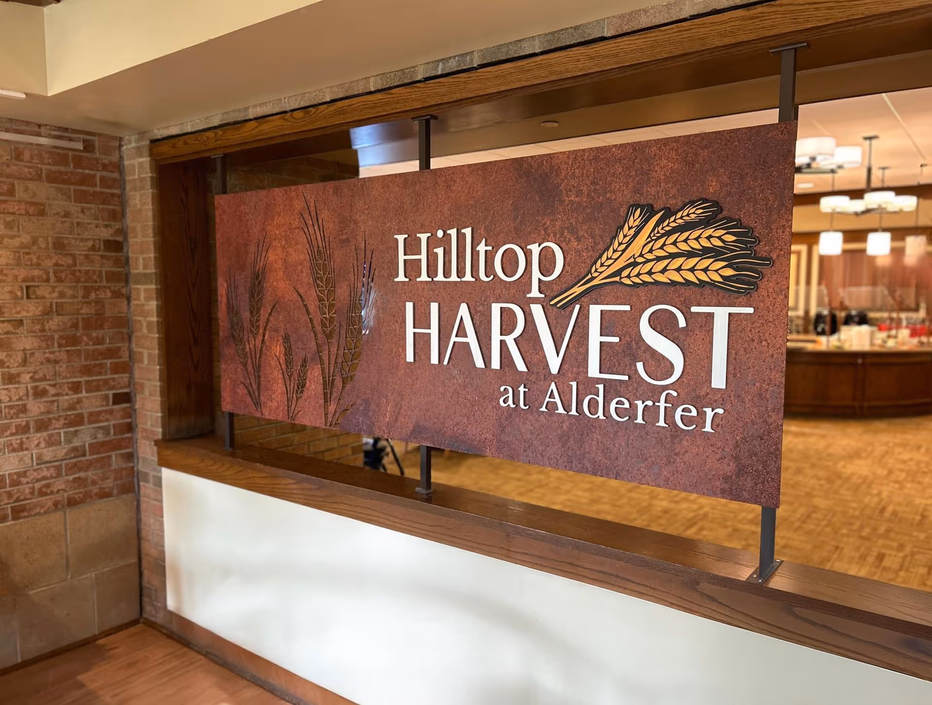 A decorative sign reading 'Hilltop HARVEST at Alderfer' with an image of wheat stalks, mounted on a wooden frame inside a building with brick and wood interior elements.