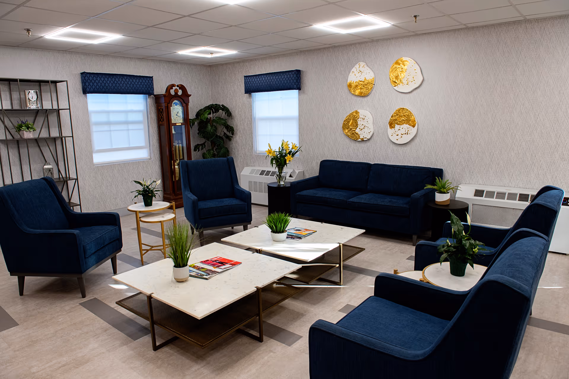 A well-lit living room with modern decor featuring navy blue upholstered sofas and armchairs arranged around two white marble coffee tables. The room has two windows with blue valances, a tall wooden grandfather clock, a metal shelving unit with decorative items, and several potted plants. On the wall, there are four decorative art pieces with gold and white accents.