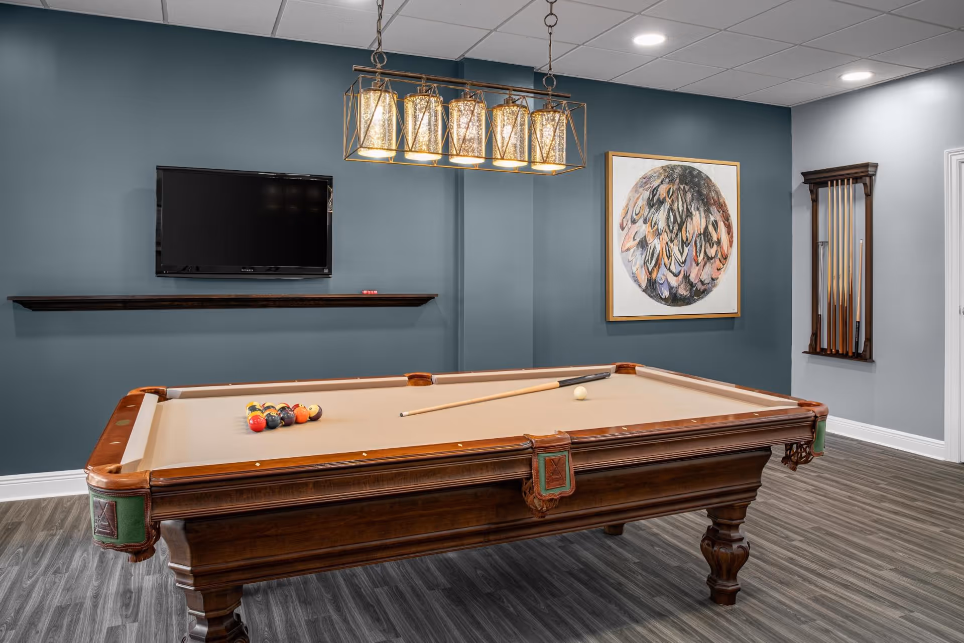A game room with a wooden pool table set up for a game, a cue stick resting on the table, a wall-mounted TV, a rack holding pool cues, a modern chandelier with five lights, and a framed abstract artwork on a blue-gray wall.