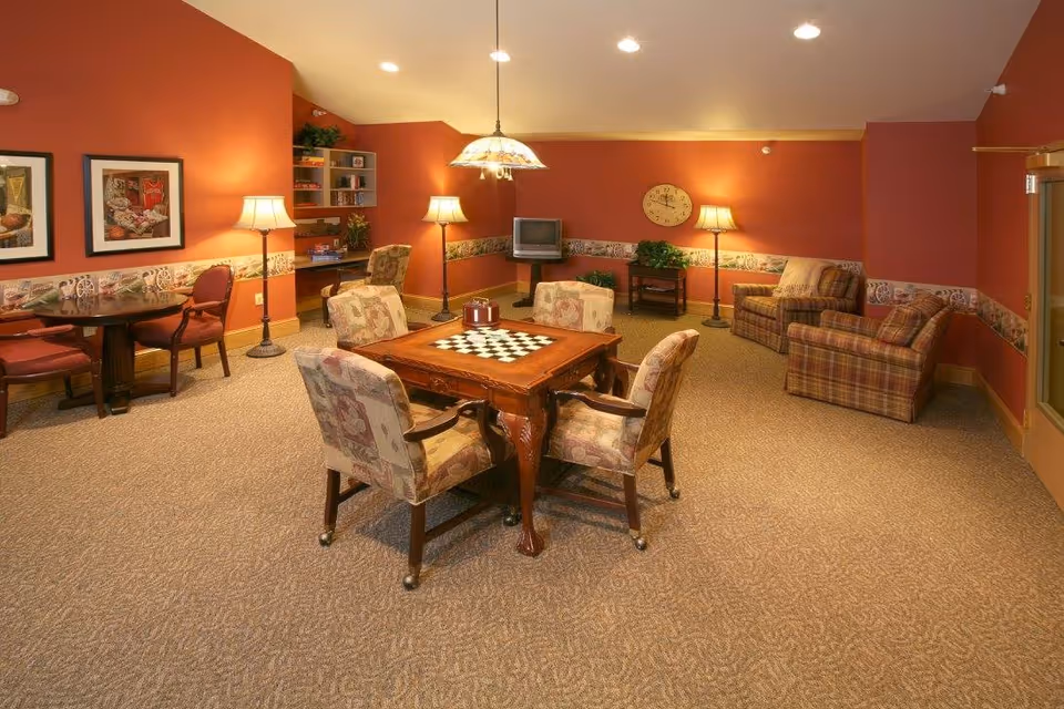 A cozy common room with warm orange walls and carpeted floor. The room features a wooden table with a built-in chessboard surrounded by four cushioned chairs. There are two plaid armchairs near the back wall, a small TV on a stand, a clock on the wall, and several floor lamps providing soft lighting. A round table with two chairs is placed against the left wall, which is decorated with framed pictures and a wallpaper border.