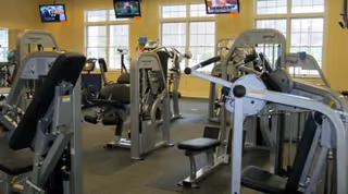 Interior view of a fitness center with various exercise machines including weight machines and cardio equipment. The room has large windows letting in natural light and three televisions mounted on the wall above the windows.