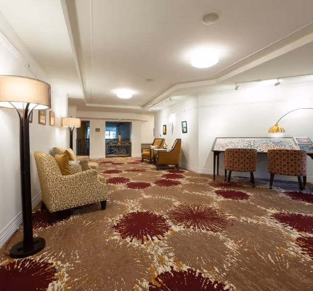 A well-lit interior common area with patterned carpet featuring large red and beige floral designs. The space includes several upholstered armchairs with cushions, floor lamps, and a table with two patterned chairs and a lamp. The walls are white with framed artwork, and the ceiling has recessed lighting.