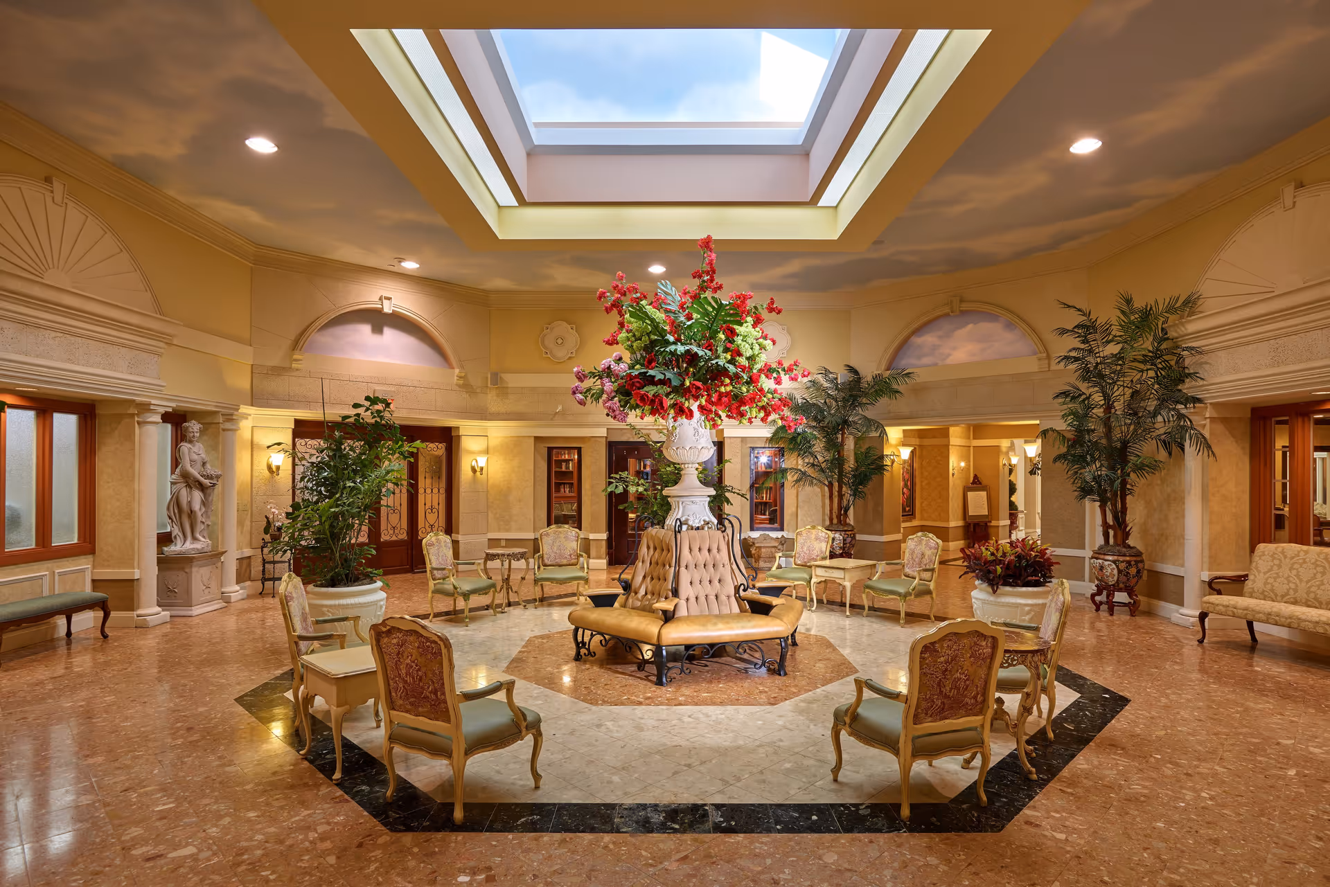Elegant circular seating area in a spacious room with a large floral arrangement in the center, surrounded by vintage-style chairs and small tables. The room features a skylight ceiling, marble flooring, classical statues, potted plants, and warm lighting, creating a luxurious and inviting atmosphere.